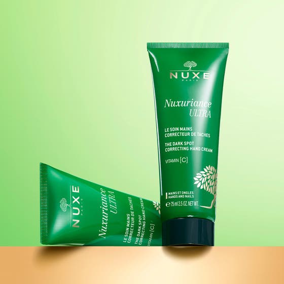 Nuxe Nuxuriance Ultra 90 g anti pigmentering handkräm visas som vit kräm på en hand bredvid den gröna tuben.