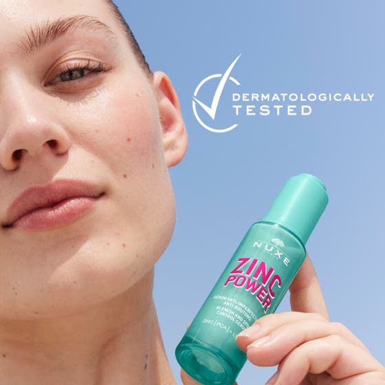 Nuxe Zinc Power Blemish And Spot Control Serum 30 ml mot orenheter i grön flaska som hålls av en person mot blå himmel