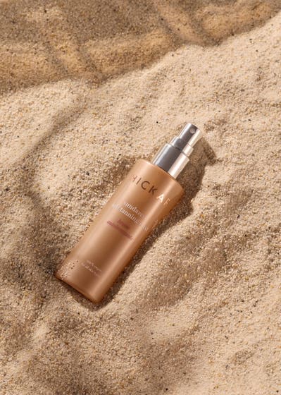 Hickap Sundazed Self Tanning Mist 100 ml brun utan sol spray i bronsfärgad flaska placerad på finkornig sand